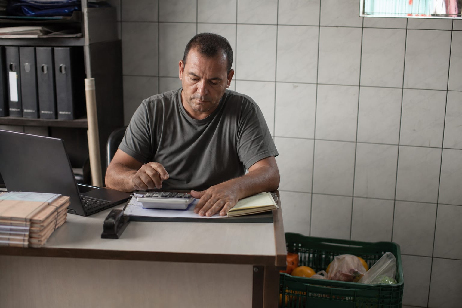 Dono de mercadinho faz contas na calculadora com caixa de produtos retirados da venda sobre a mesa